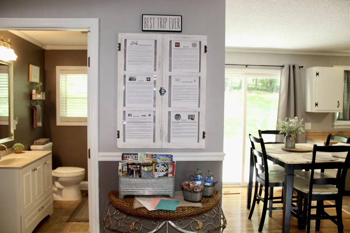 A cozy dining area with a round table set for four, a decorative sign reading 'BEST TRIP EVER', and a wall-mounted information board. The space is staged for family meals, planning outings, or relaxing with friends, exuding comfort and a sense of gathering.
