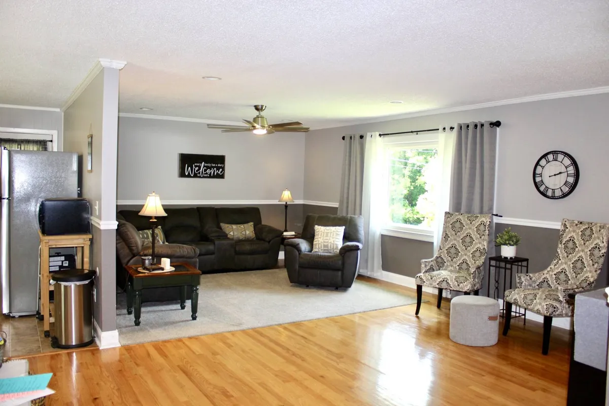 The image showcases a bright, inviting living room with a plush sofa set arranged near a large window that fills the space with natural light. A charming welcome sign and a stylish clock add warmth and personality, hinting at a cozy atmosphere perfect for gatherings or quiet moments.