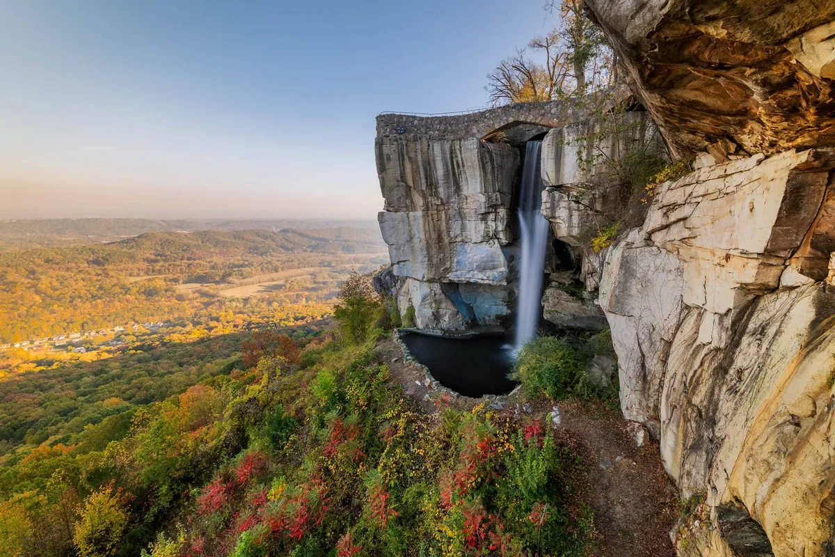 A scenic view of a cascading waterfall surrounded by rock formations and lush greenery, opening up to a tranquil valley below. The image conveys a sense of nearby natural beauty and opportunities for outdoor exploration.