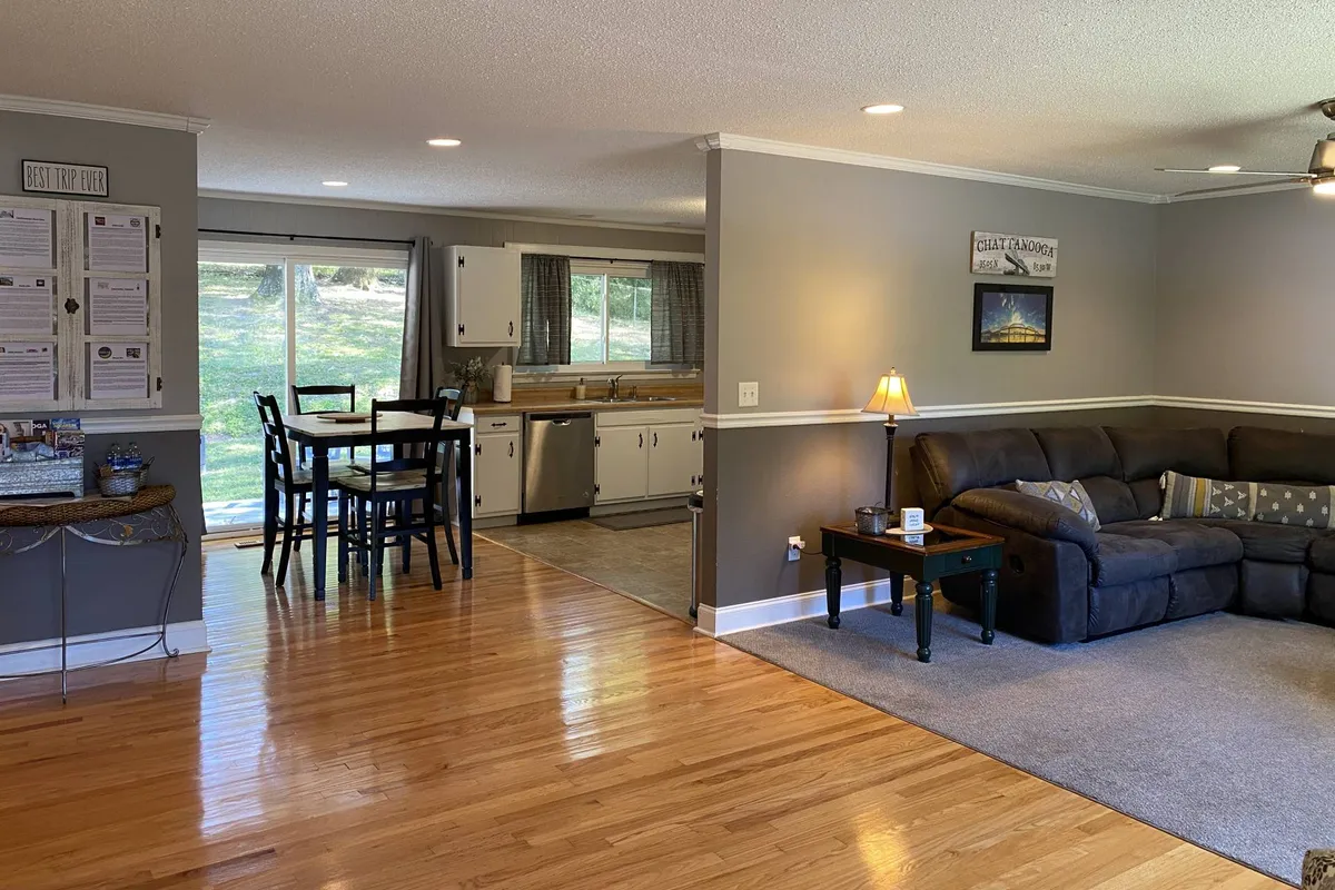 A bright, open-plan living and dining area with a dining table, comfortable sofa, and kitchen area all bathed in natural light from large windows. The scene captures a relaxed, family-friendly atmosphere suitable for gatherings, casual dining, or watching movies together.