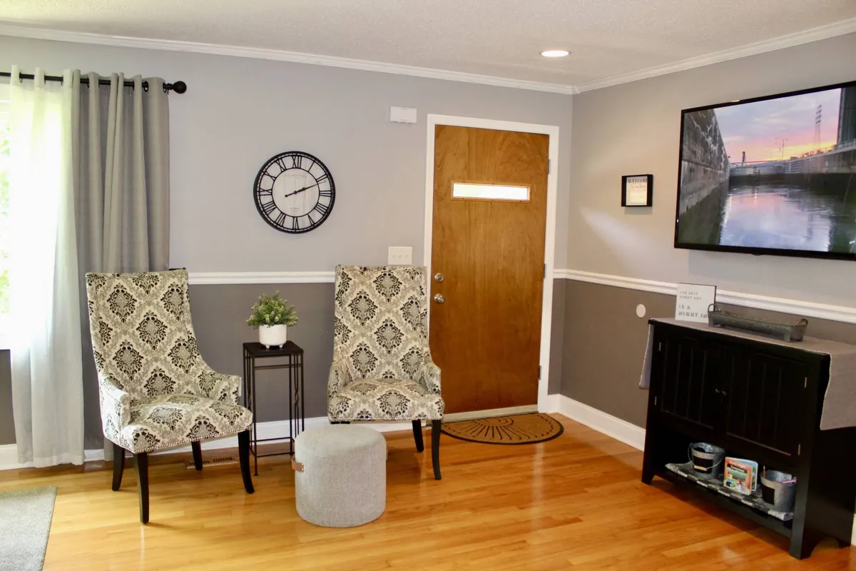 A stylish interior space featuring two patterned chairs beside a wooden door, a television displaying a scenic image, and a small side table. The arrangement invites socializing, relaxation, and cozy moments indoors.