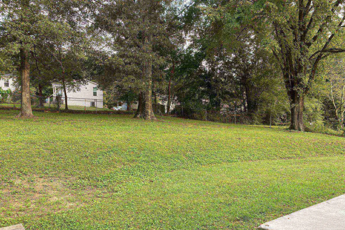 A perspective of the backyard with lush green grass, tall trees, and a visible fenced area enclosing the space. The white house sits in the background, underscoring the peaceful, private setting and creating a safe environment for children and pets to play freely.
