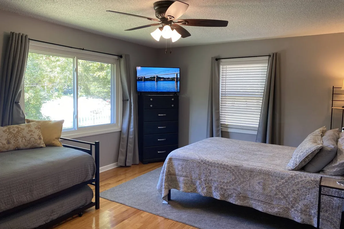 A serene and comfortable bedroom featuring a large bed with decorative pillows, a large window that fills the room with natural sunlight, a Smart TV displaying a scenic image, and a dresser. The space looks ideal for unwinding, with plenty of room and a soothing palette.