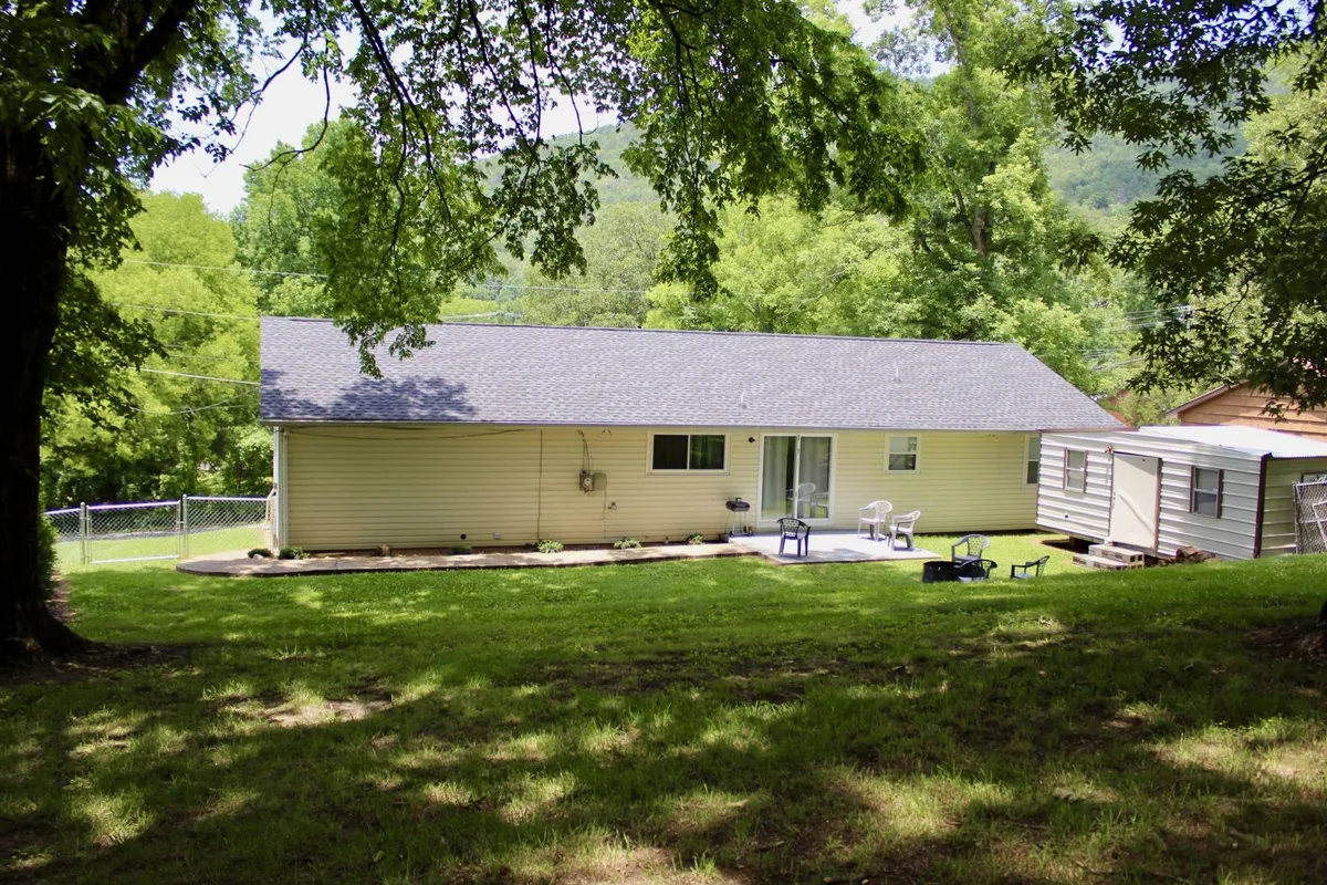 A wide view of the backyard featuring a lush green lawn, spacious patio area, and mature trees surrounding the space. The patio includes outdoor seating, providing a comfortable spot to relax or gather with family and friends. The peaceful environment is highlighted by natural sunlight and ample room to play or entertain.
