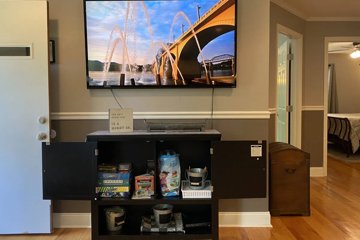 A living room corner featuring a TV displaying a scenic view and a storage cabinet filled with games and assorted items. The space is designed for entertainment and leisure, inviting families to enjoy movie nights or board games together.