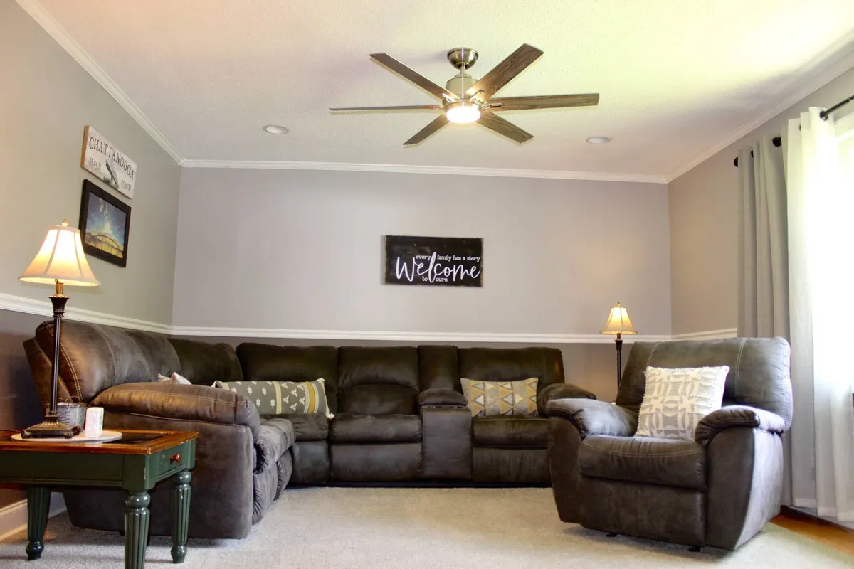 A spacious living room with a comfortable sectional sofa adorned with decorative pillows. The room also features a welcome sign, warm lighting from lamps, and an inviting arrangement perfect for socializing or relaxing with family. The cozy atmosphere encourages togetherness and shared activities.