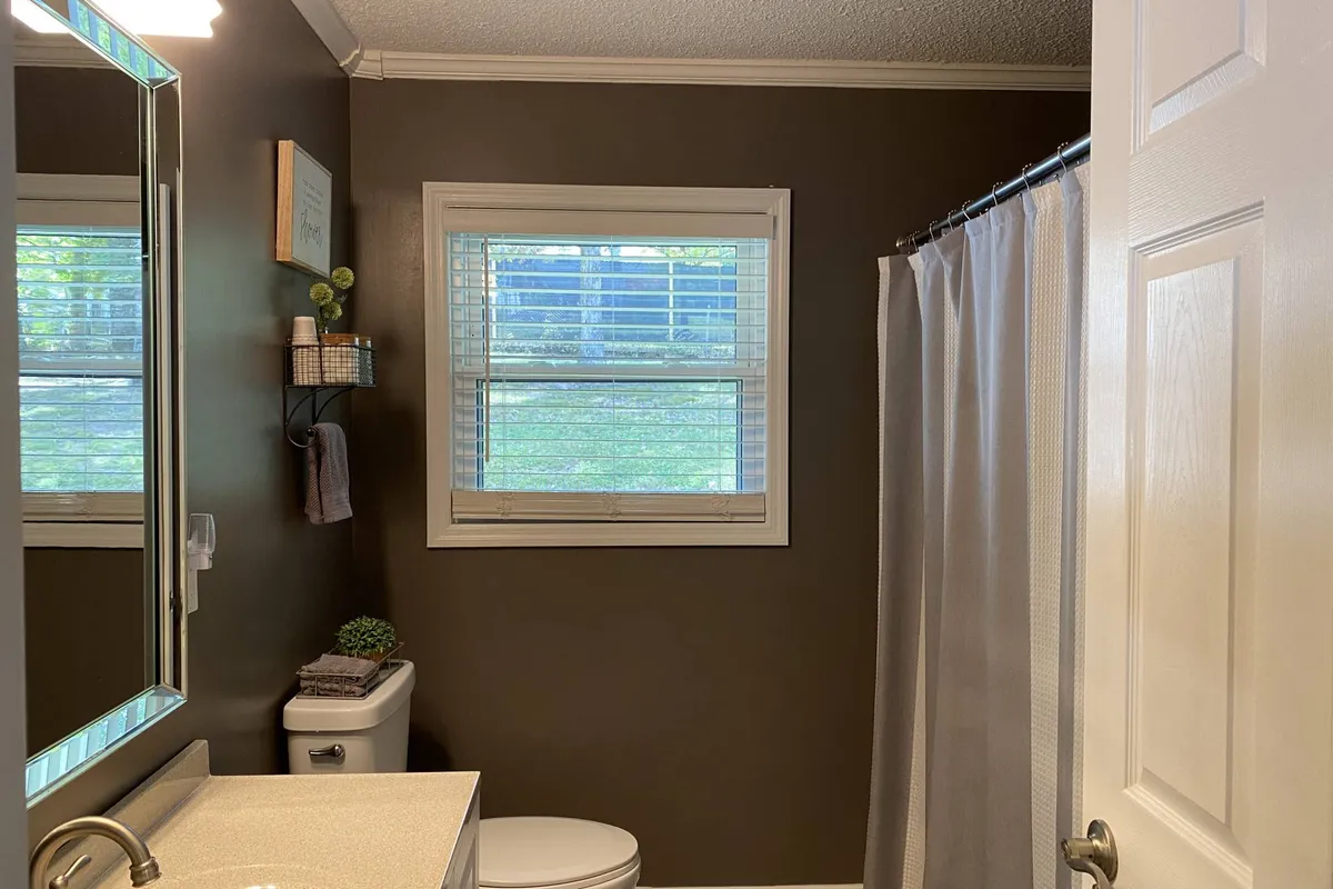 A bright and clean bathroom featuring a mirror, sink, toilet, shower curtain, and a window allowing natural light. The space looks well-stocked and serene, perfect for relaxation and self-care.